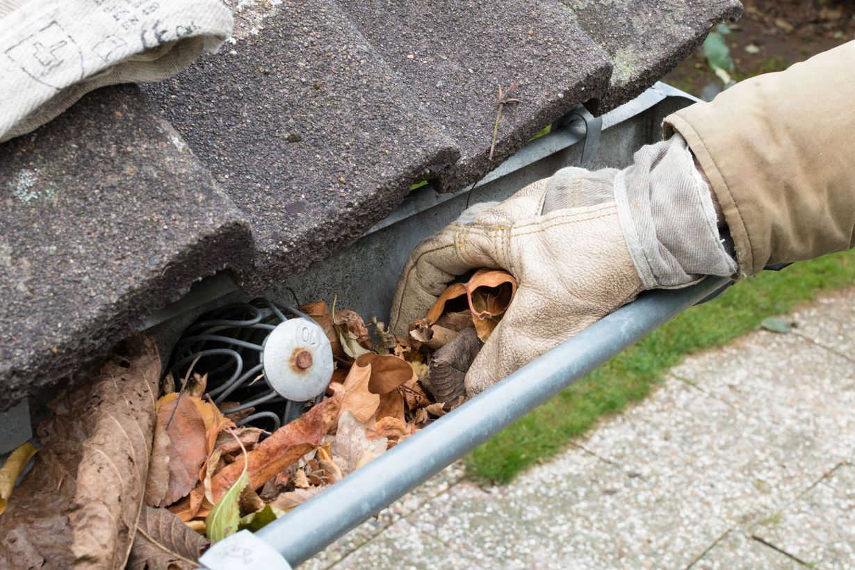 Gloved hand clearing leaves from a roof gutter with a brush and drain opening visible
