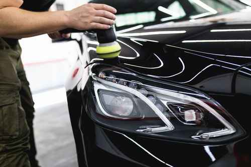 Person polishing a black car’s headlight and hood with a buffer in a garage