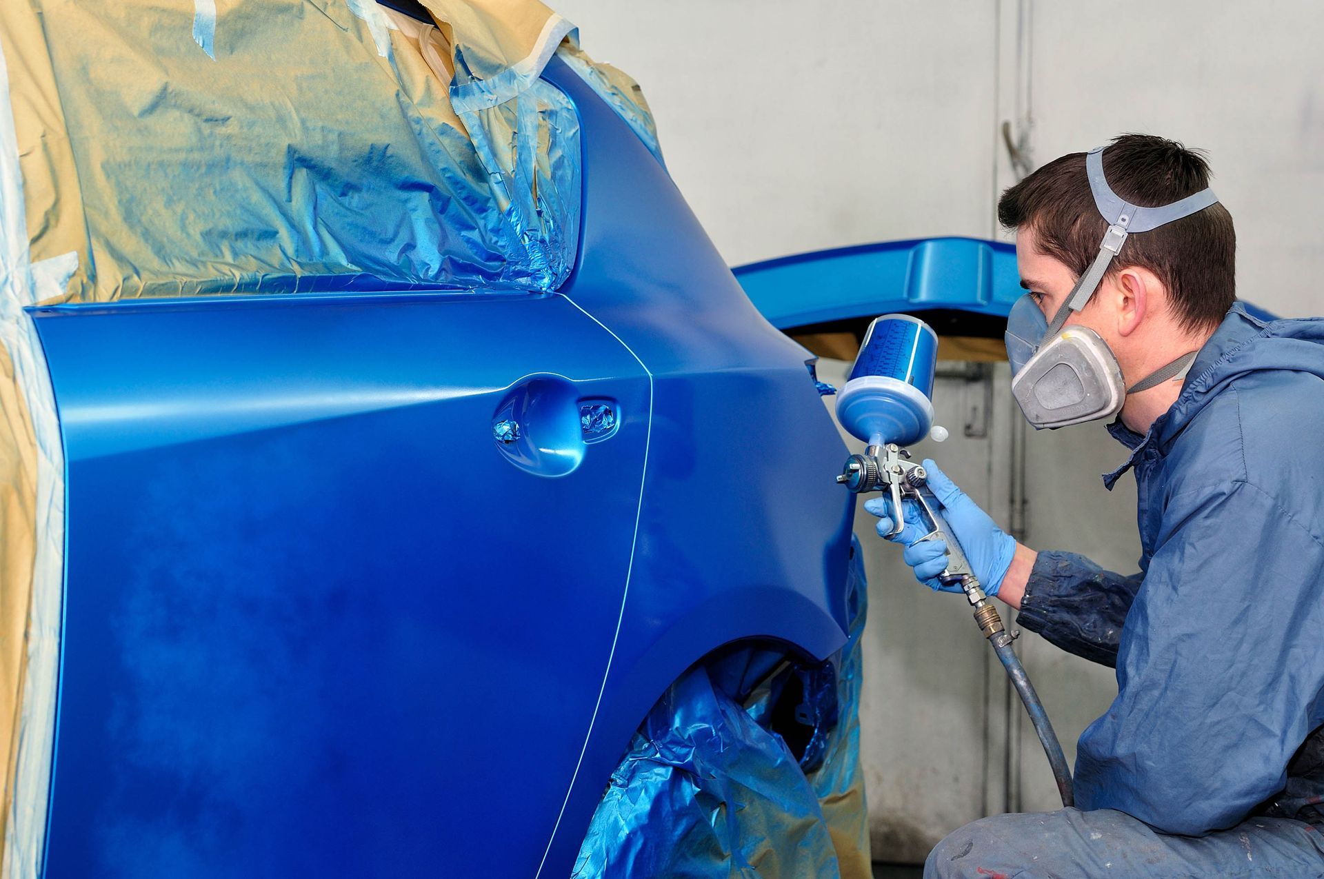 Auto body painter spraying blue paint on a car in a workshop, wearing a respirator and gloves