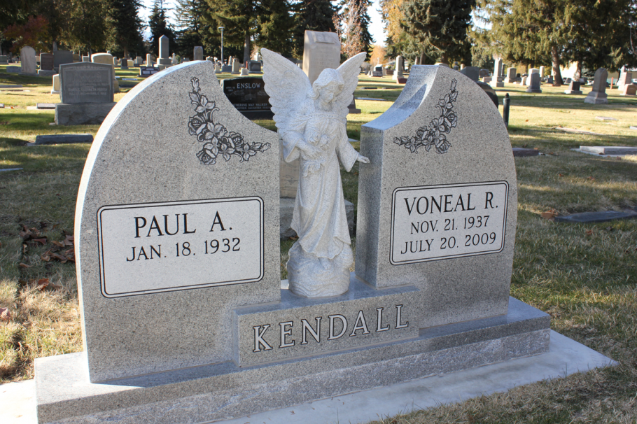 Headstone in a cemetery, gray stone with angel statue between two sections, names and dates.