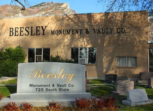 Beesley Monument & Vault Co. building and sign. Light brick, gold lettering, green shrubs, and some grave stones.