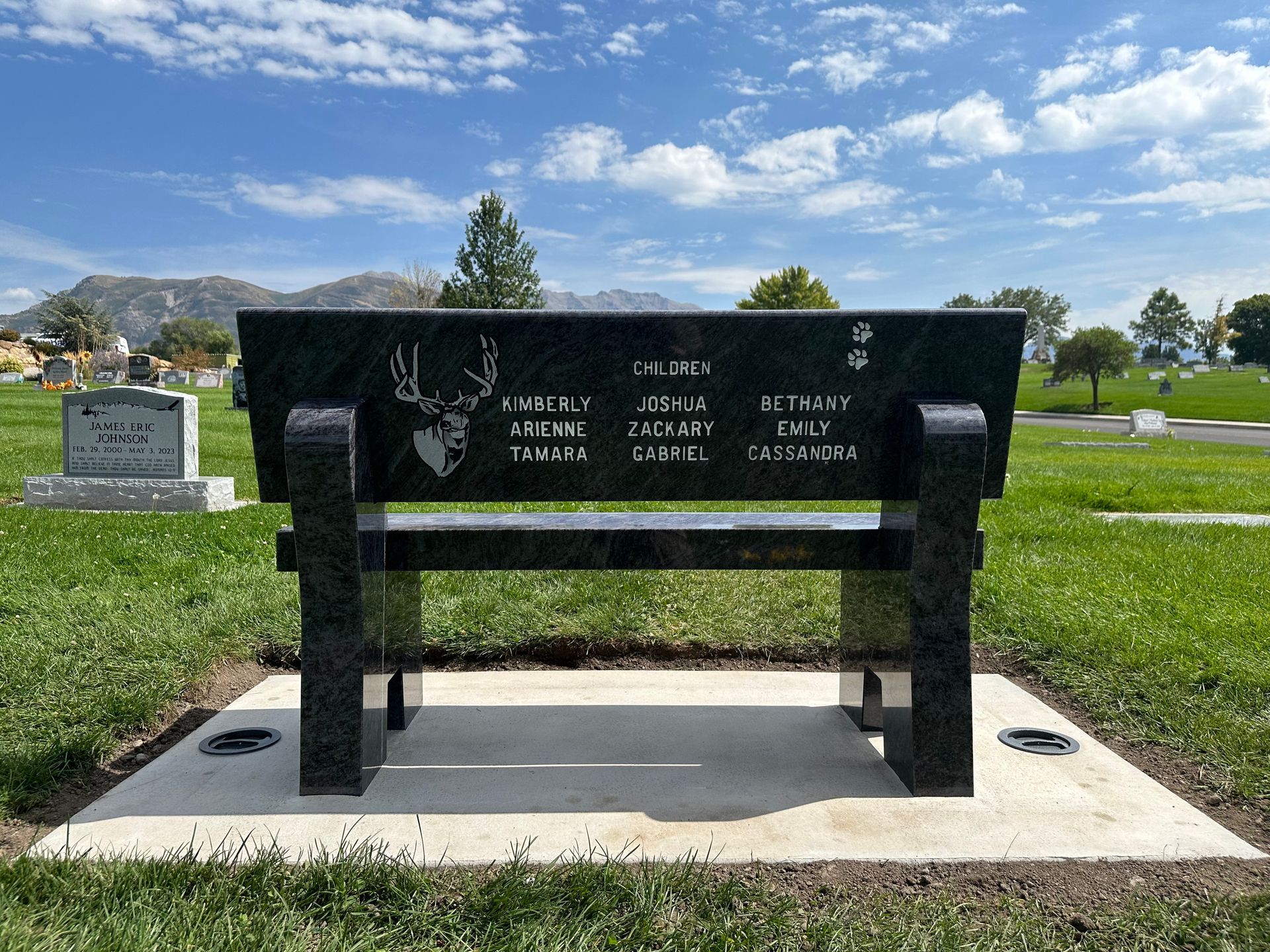 Black granite bench in a cemetery with names and a deer head etched. Mountain backdrop.