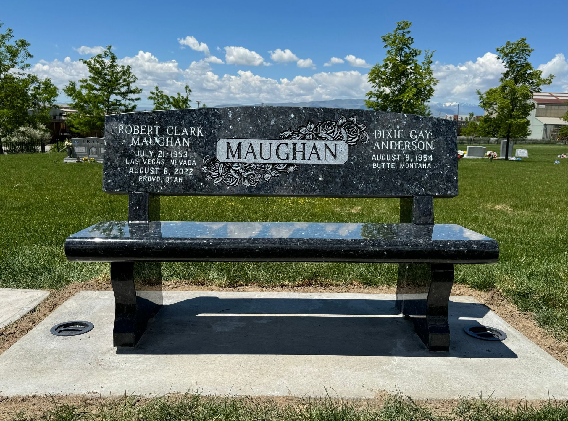 Black granite bench with Maughan and names engraved, set in a green park under a blue sky.