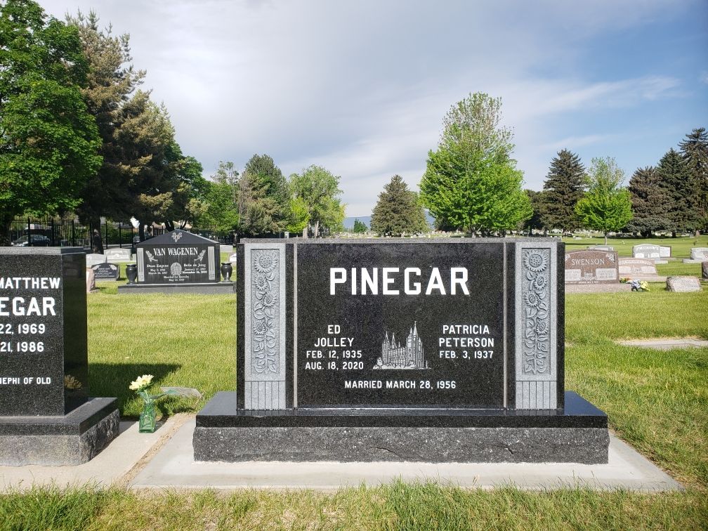 Black headstone in a cemetery; Pinegar is inscribed, with names and dates. Green grass, trees, and blue sky.