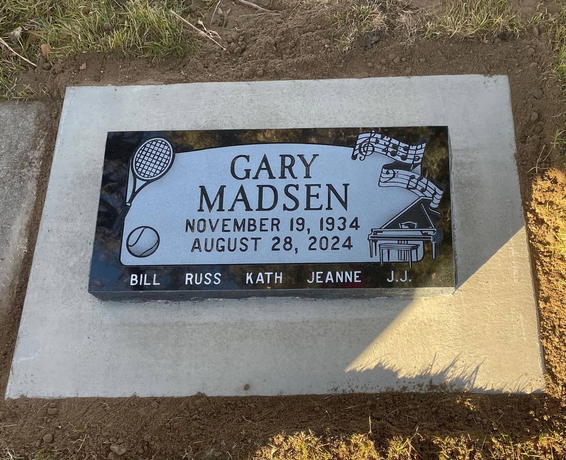 Grave marker for Gary Madsen, with tennis racket and piano illustrations, dates Nov 19, 1934 - Aug 28, 2024.