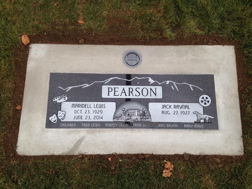 Tombstone of Pearson with names, dates, and etched imagery: drama masks, film reel, figures, mountain range.