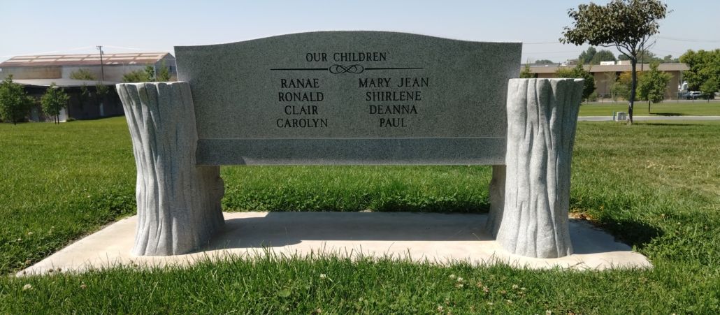 Gravestone in the shape of a bench with names inscribed; located in a green grassy area.