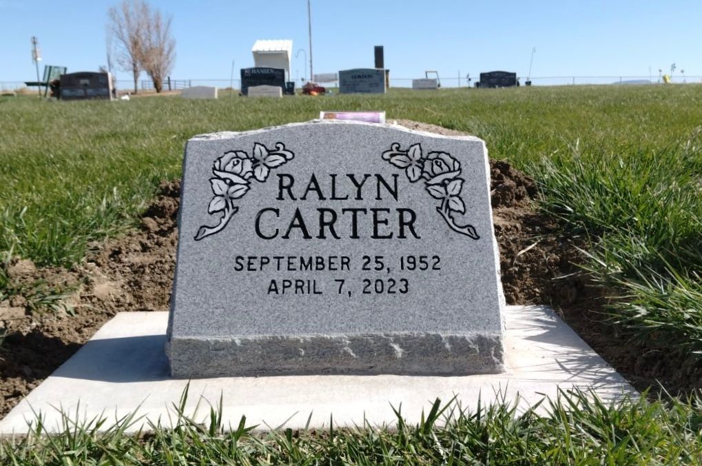 Headstone for Ralyn Carter, born 1952, died 2023, in a cemetery; gray stone, floral accents, green grass.
