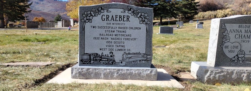 Gravestone in a cemetery, GRAILER. White with black text, grassy surroundings, mountain in the distance.