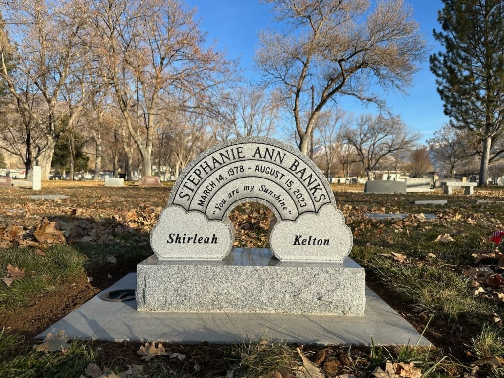 Gravestone shaped like a rainbow in a cemetery. The names Shirley & Kellon flank the clouds below.
