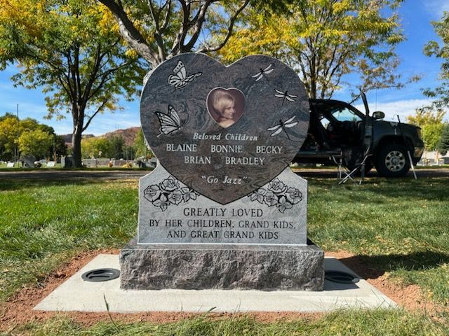 Heart-shaped tombstone in a cemetery; features names, a photo, and Greatly Loved.