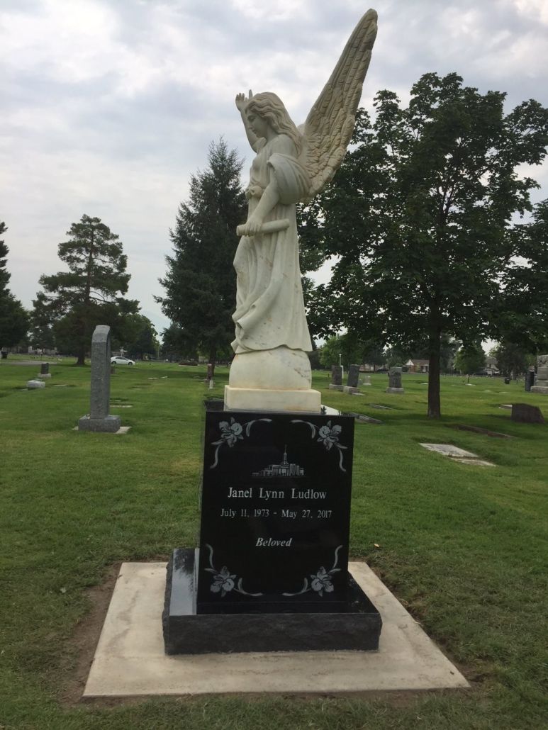 White angel statue on black tombstone in a cemetery, with names and dates.