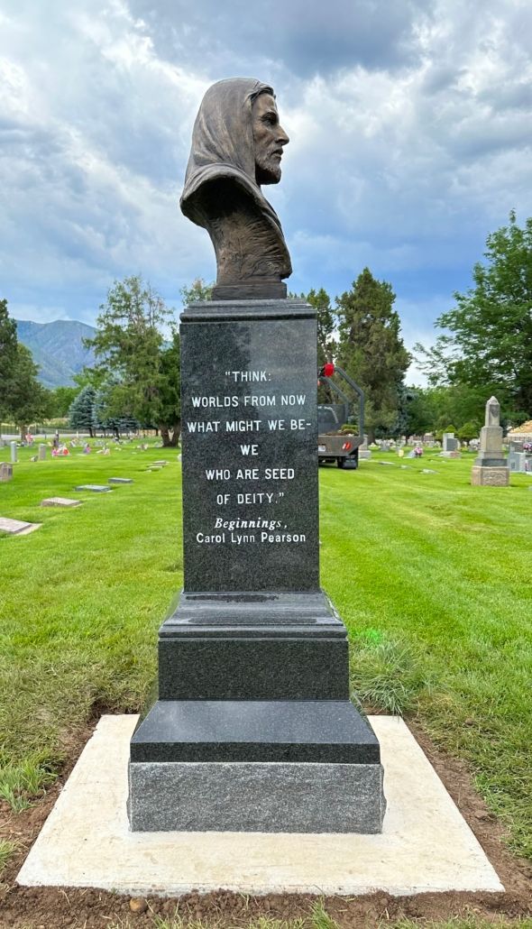 Bronze bust on black stone pedestal in a cemetery. Mountains in background, grass and headstones surrounding.