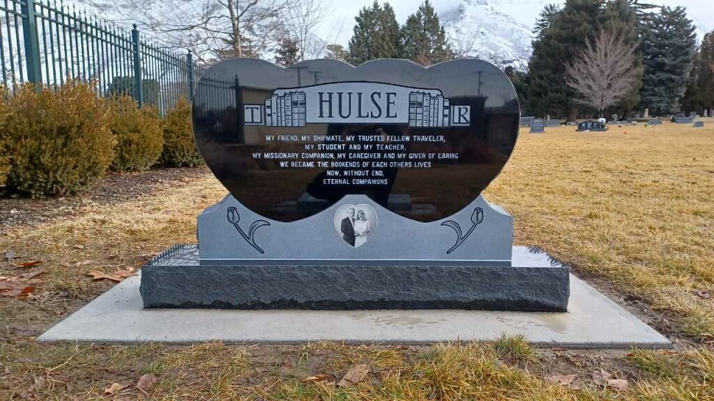 Heart-shaped black granite headstone with inscription, in a cemetery setting; mountains in the background.