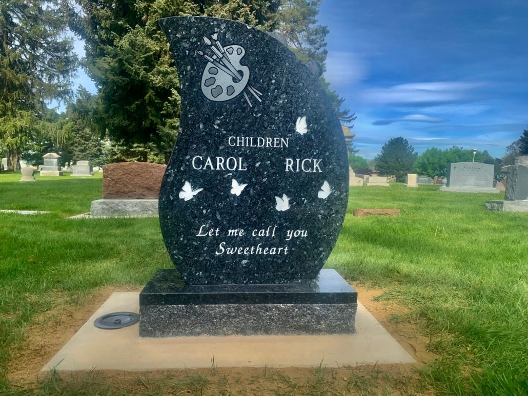 Black granite headstone with art palette, doves, names Carol & Rick, in a cemetery.