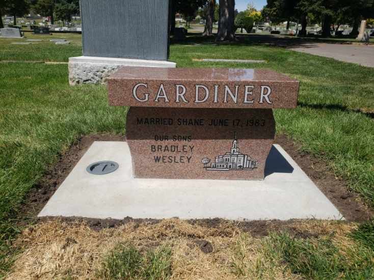 Red granite headstone engraved with GARDINER, dates, and a building silhouette; set in a cemetery.