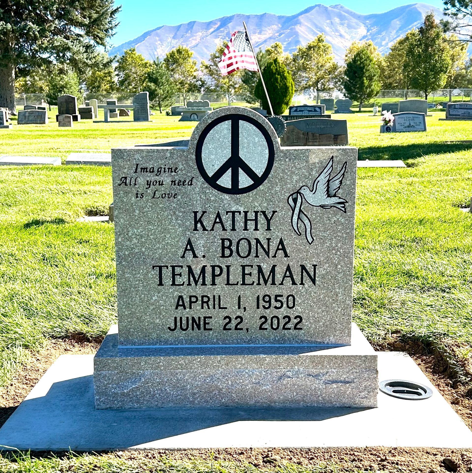 Gravestone of Kathy A. Bona Templeman in a cemetery, with a peace sign, dove, and U.S. flag; mountains in the background.