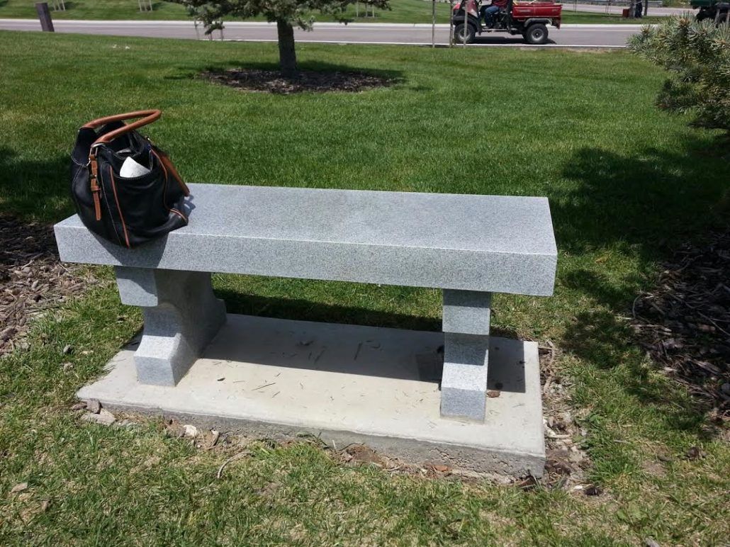 A gray granite bench with a black bag on it sits on grass, with a red vehicle in the background.
