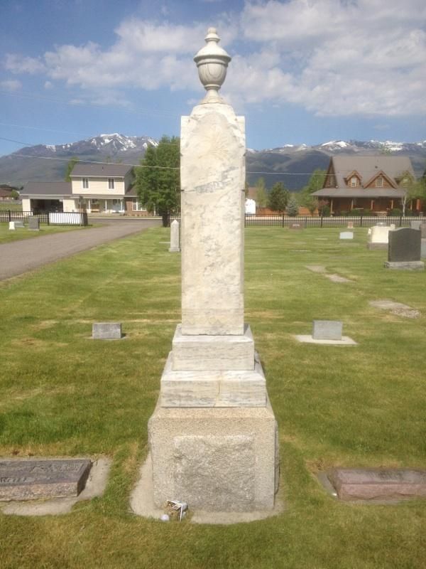 Tall white marble grave monument in a grassy cemetery, with mountains and buildings in the background.