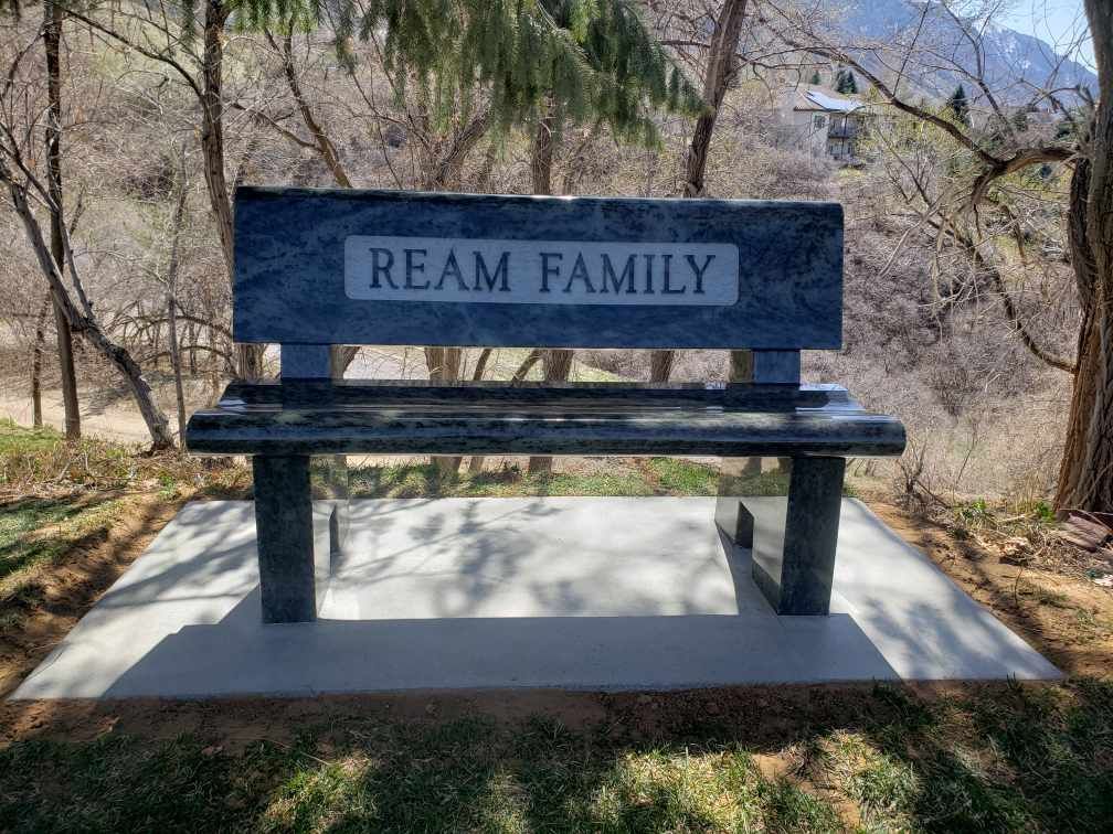 Stone bench with REAM FAMILY engraved on the back, on a concrete pad, in a park-like setting.