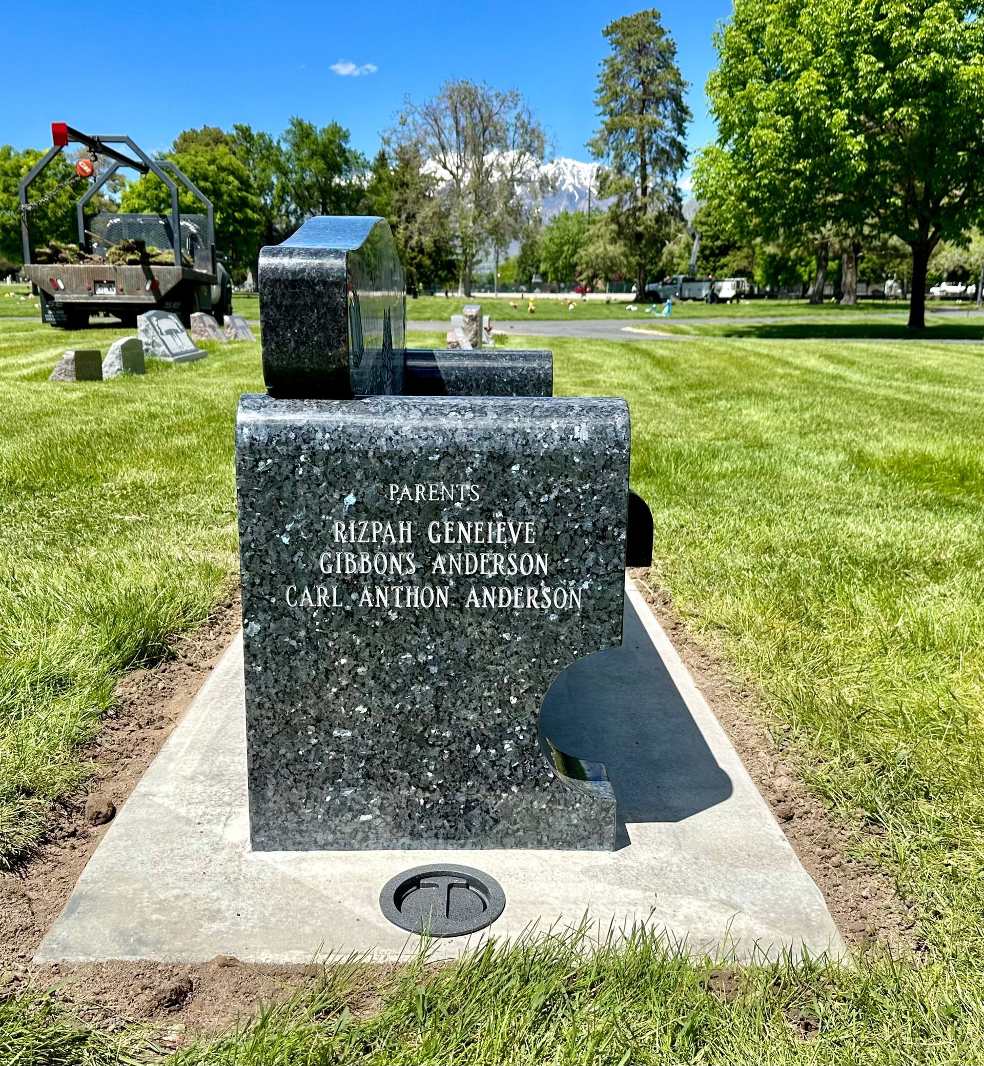 Granite headstone in cemetery with names inscribed, surrounded by grass and trees.