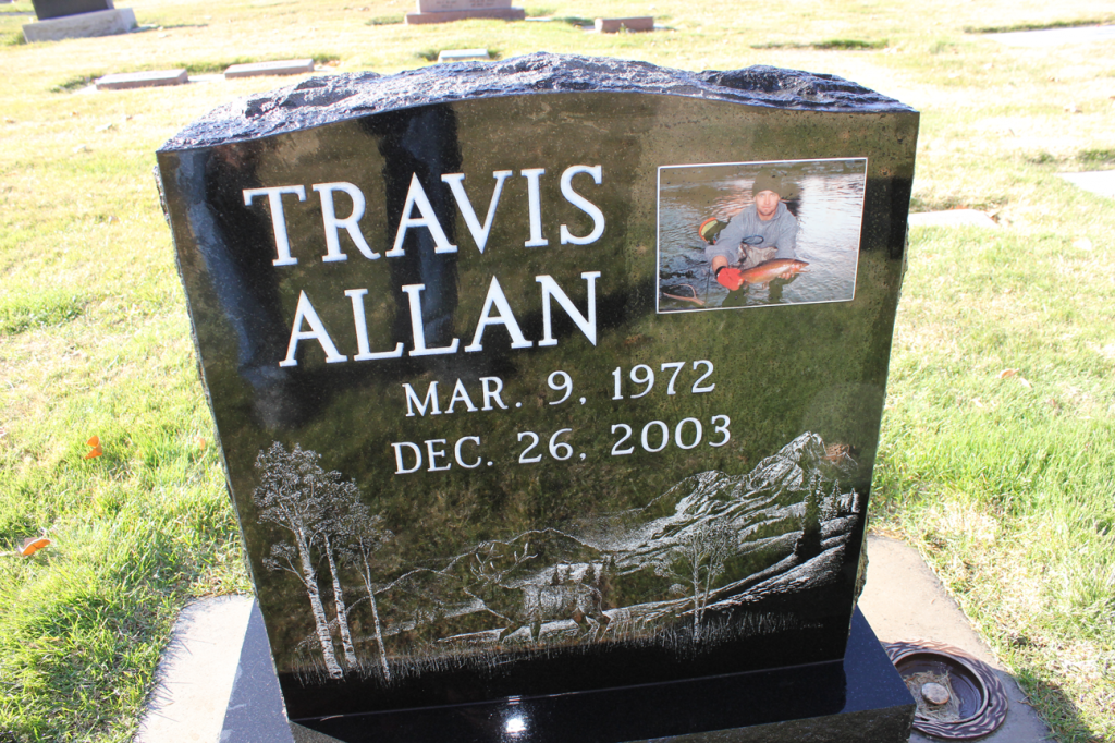 Black headstone with Travis Allan, dates, a photo, and etched landscape.