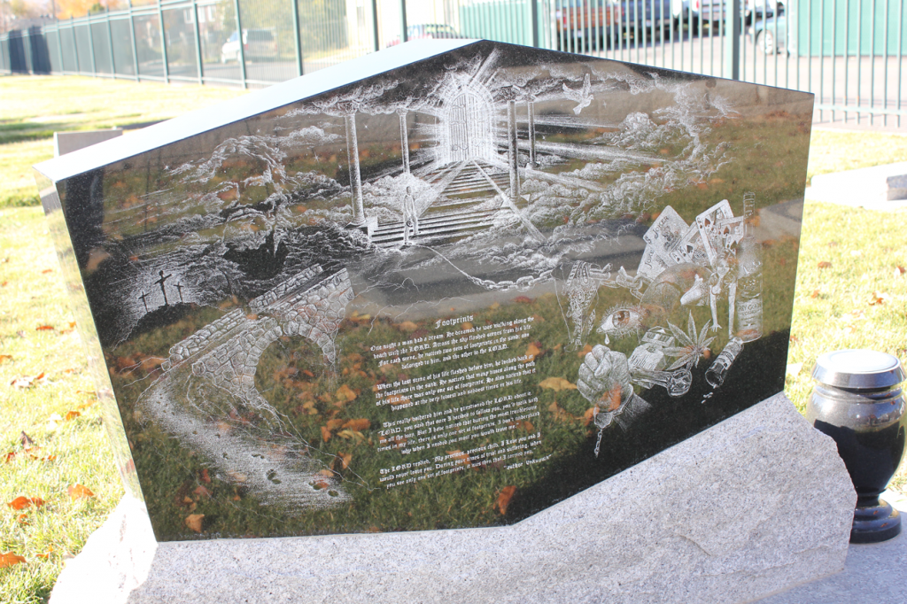 Headstone with etched river scene and inscription on a grassy field, black vase to the right.