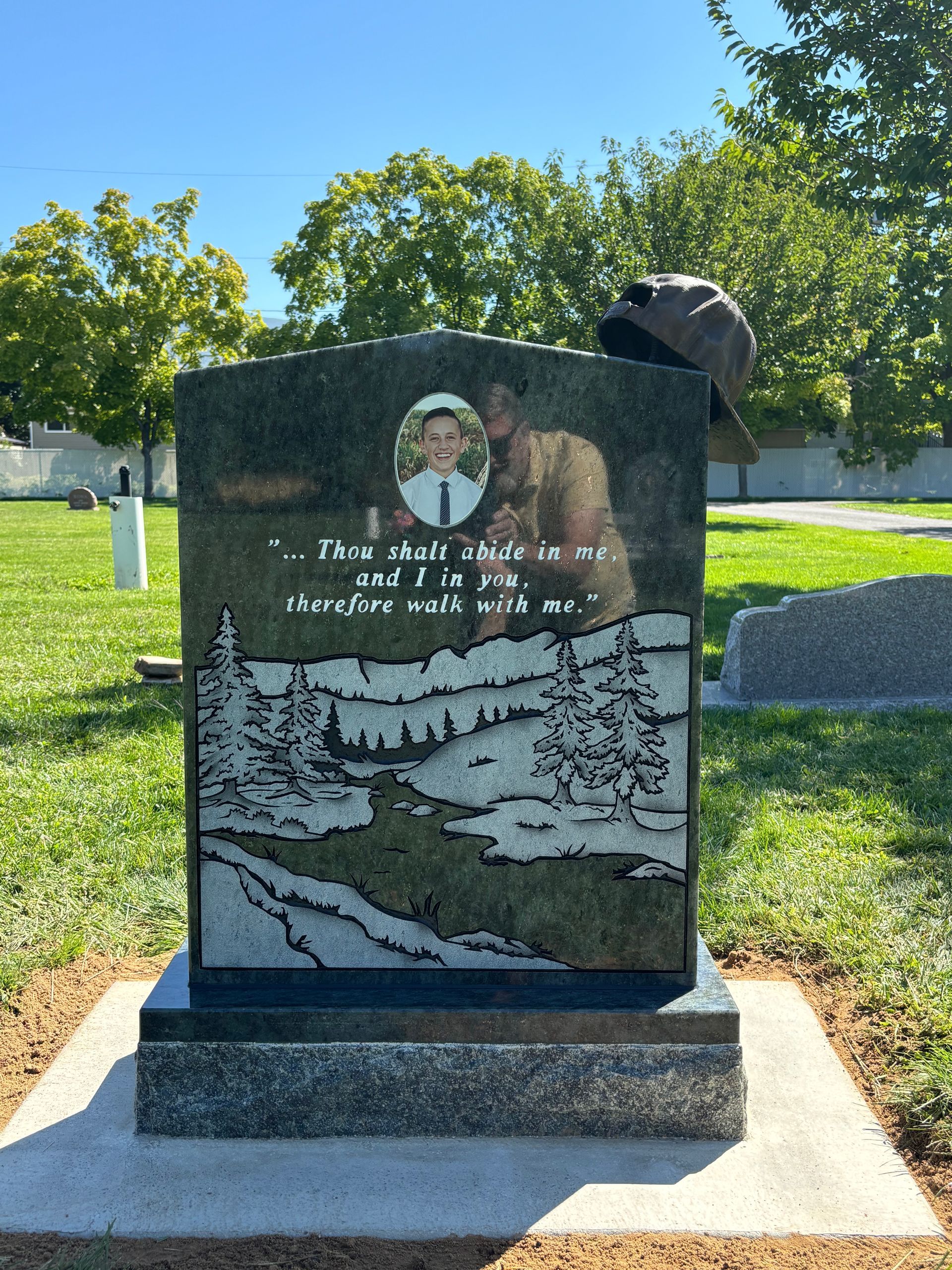 A granite tombstone with a child's portrait and landscape etching. A hat rests on the top.