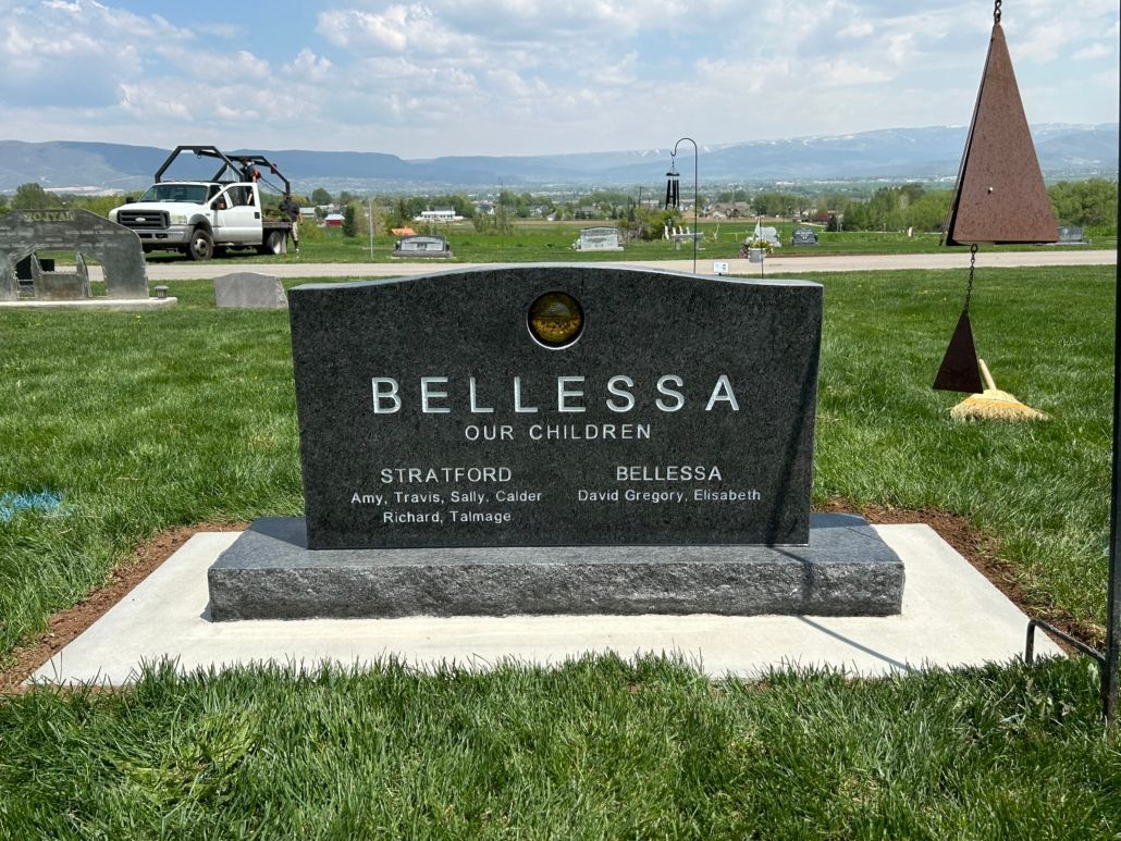 Headstone in a cemetery, reads BELLESSA Our Children. Green grass, blue sky, and mountains in the background.
