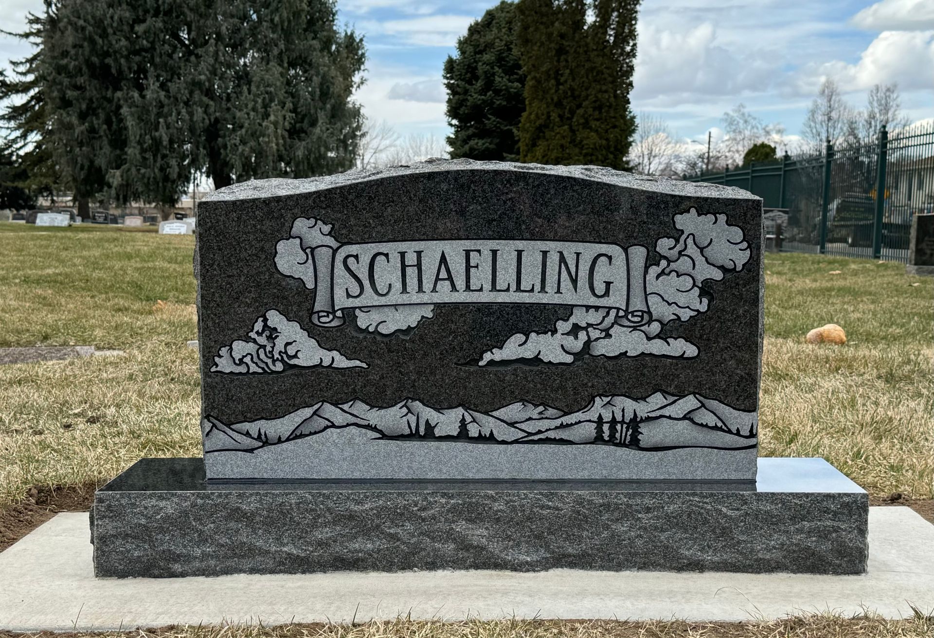 Headstone with the name SCHAELLING engraved, featuring mountain and cloud designs.