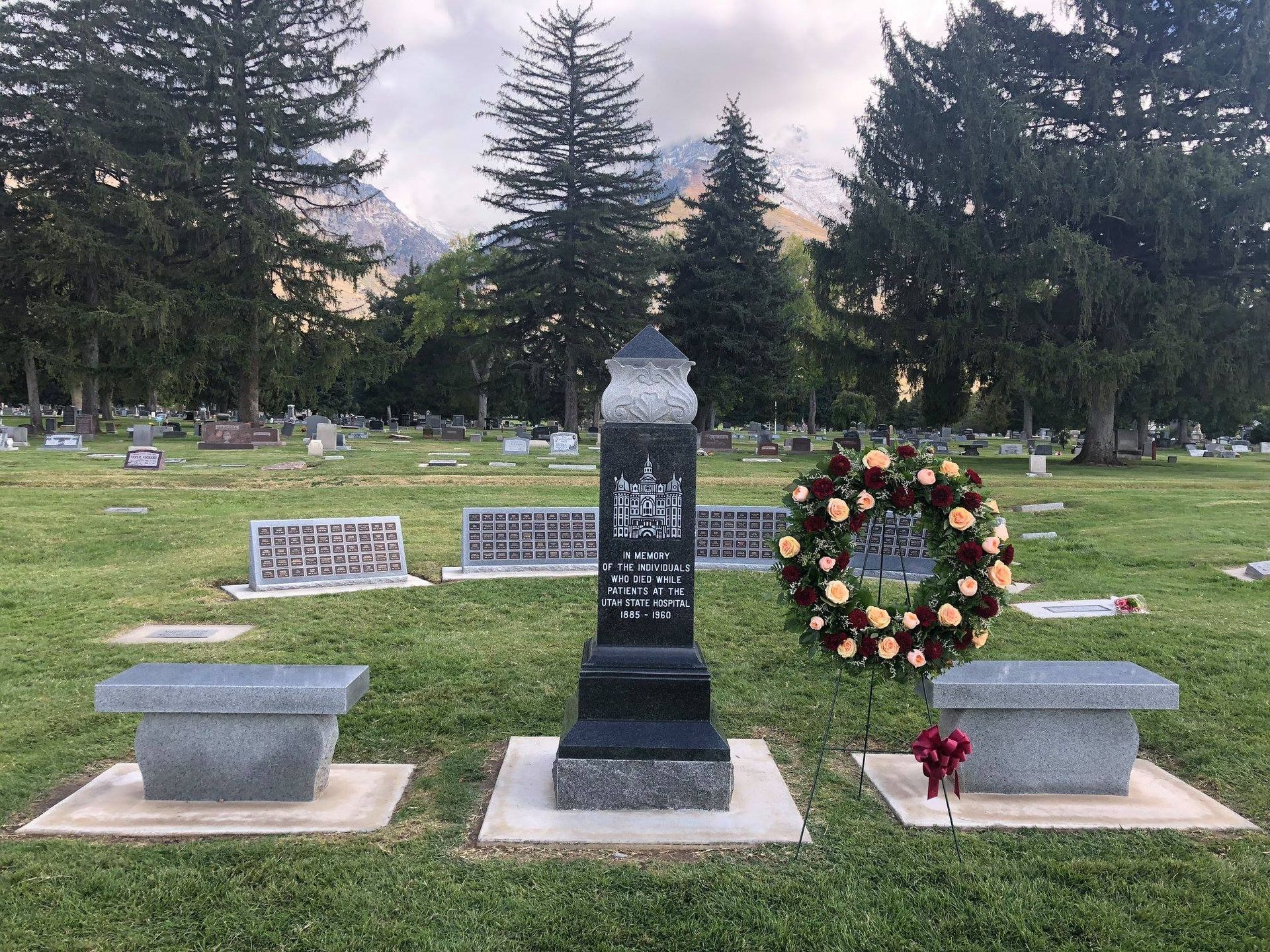 Grave with black headstone, two benches, a wreath, and a mountain backdrop in a cemetery.