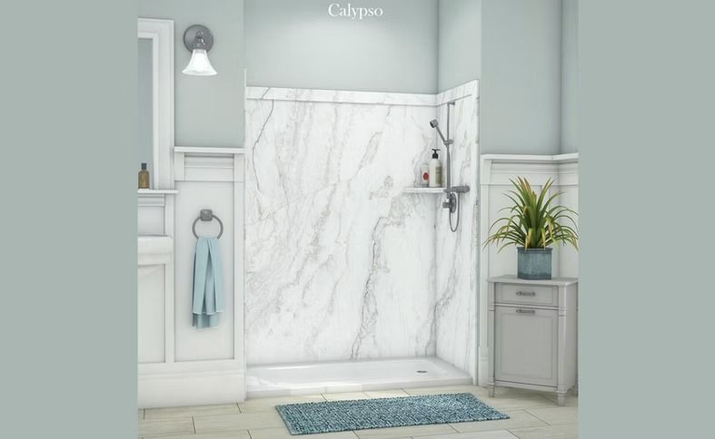 Bathroom with a white marble-patterned shower surround, a showerhead, and a small blue rug. A small cabinet and a houseplant are nearby.