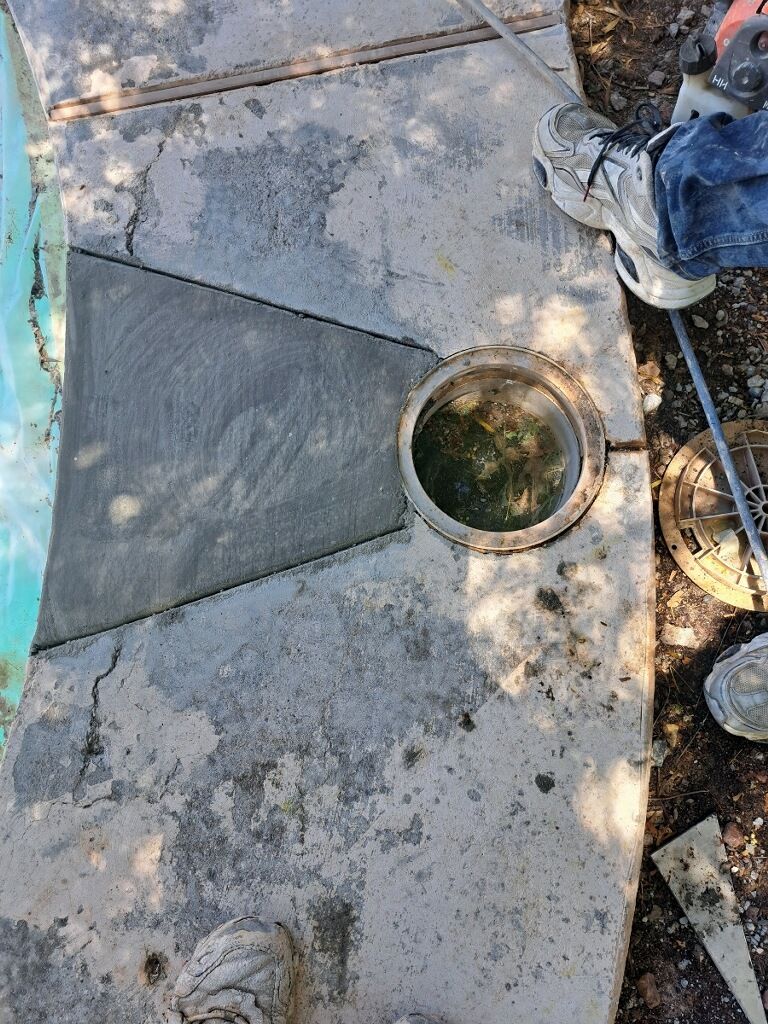 A person is standing next to a drain on a sidewalk next to a pool.