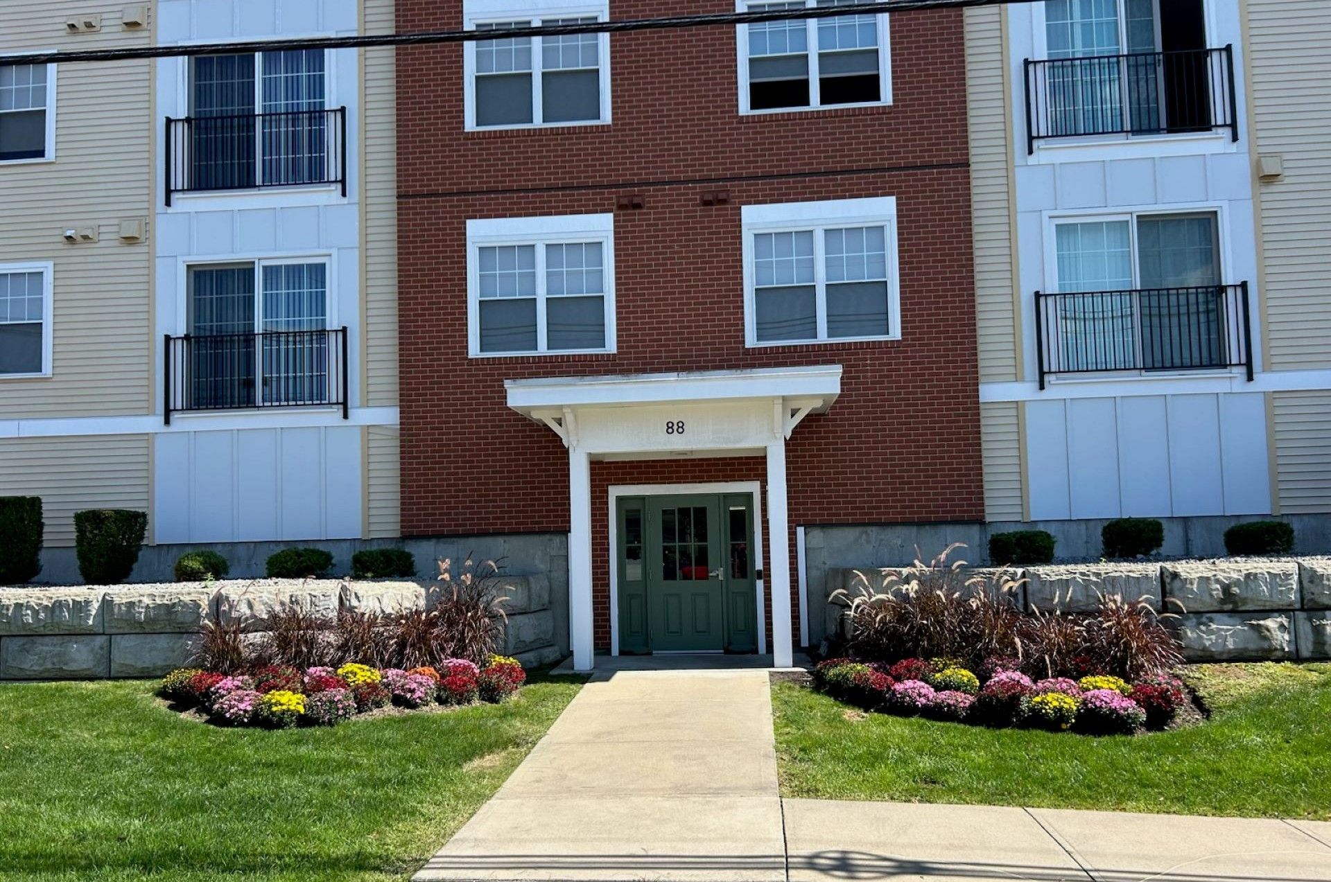 Apartment building exterior, brick and siding with small balconies, flower beds, and a green front door.