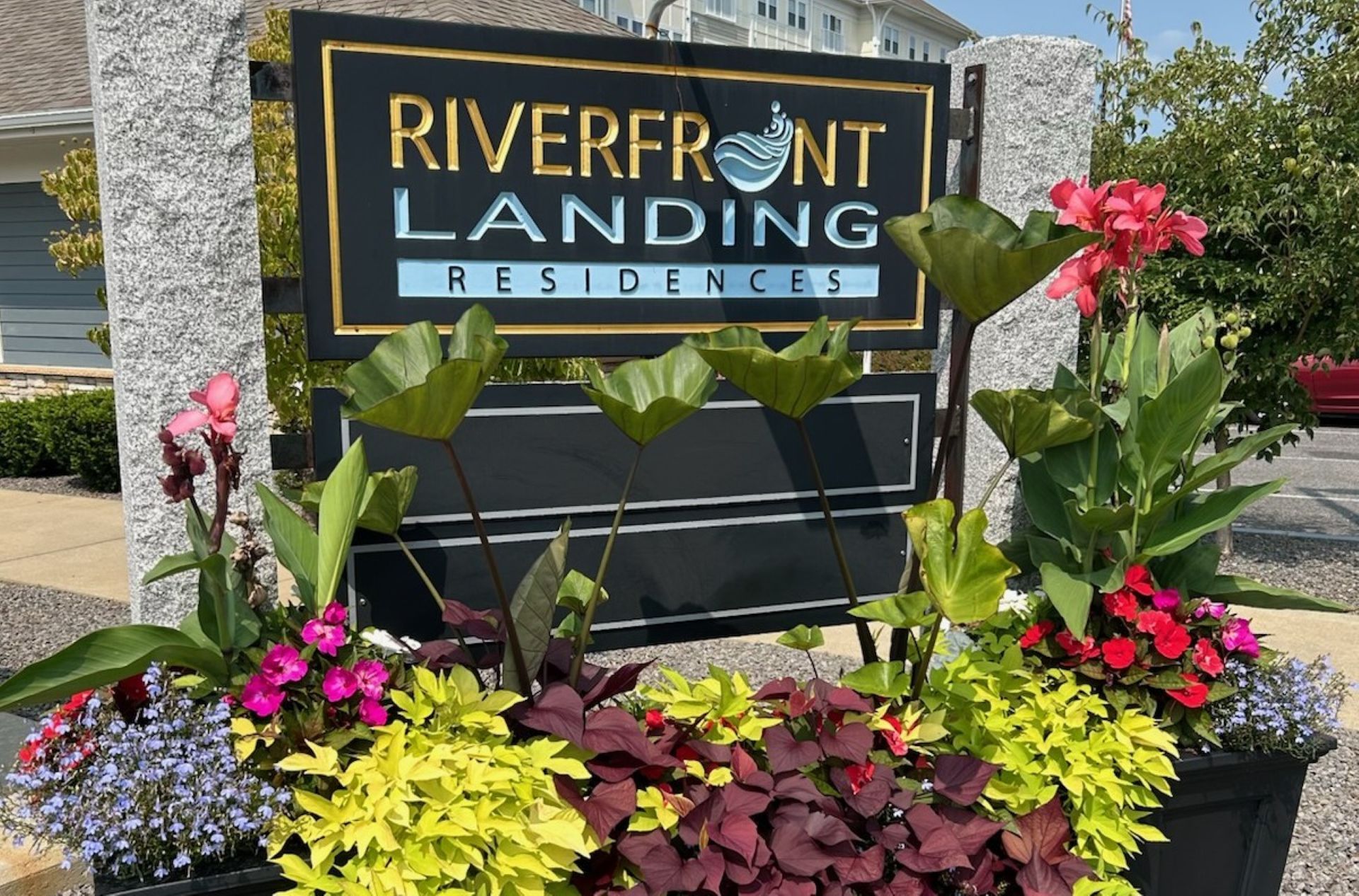 Sign for Riverfront Landing Residences, black with gold trim, surrounded by colorful flowers.