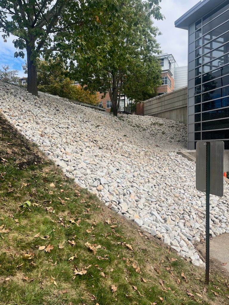 White gravel embankment beside a building, with trees and a sloped grassy edge in front.