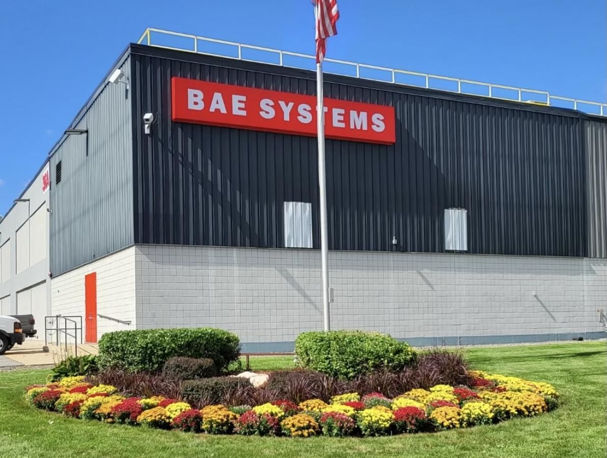 BAE Systems building with red sign, American flag, and floral landscaping.