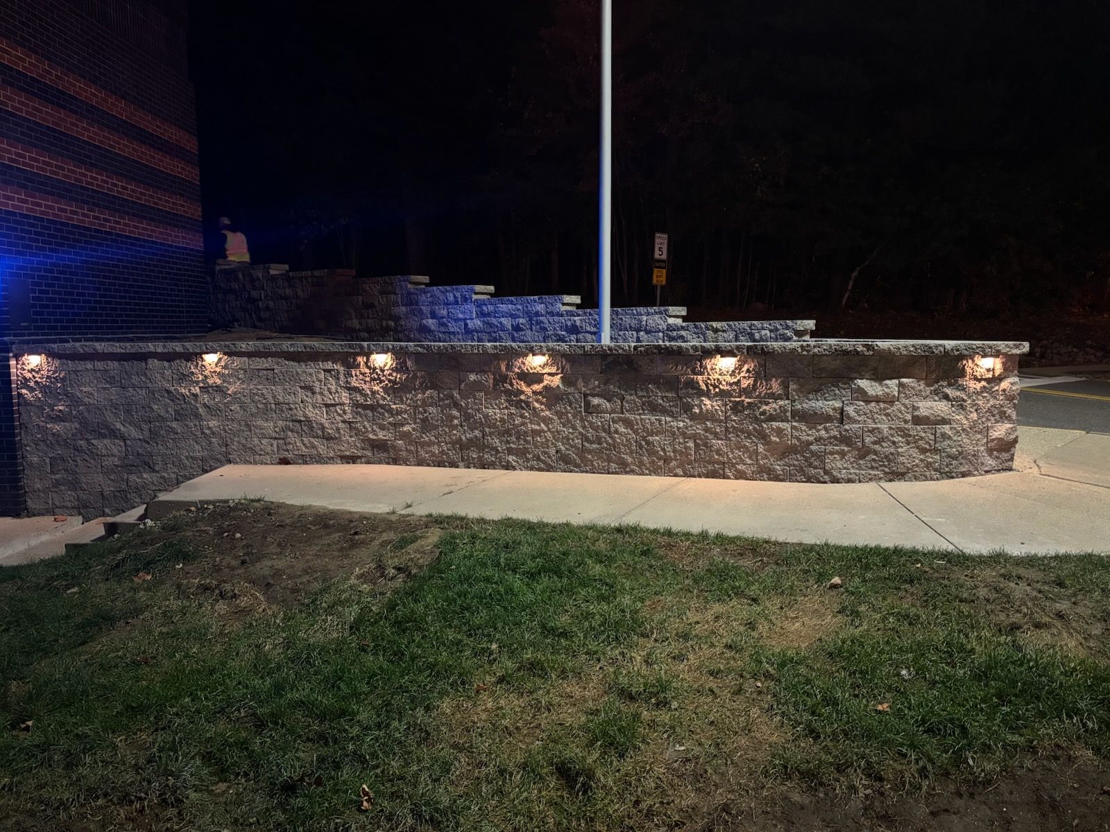 Stone wall with built-in lights at night next to grass and a building.