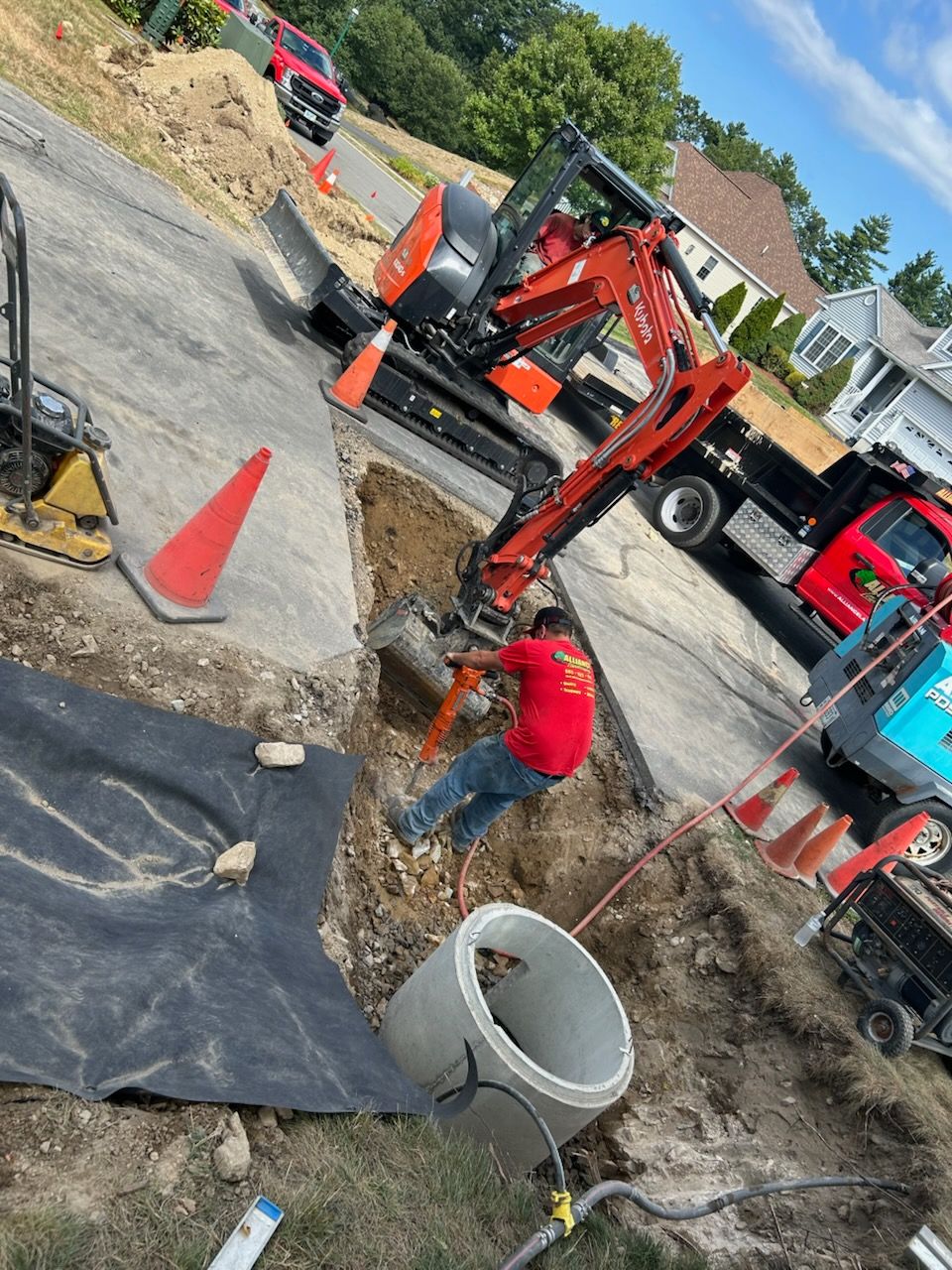 Construction worker operating equipment on a roadway, with excavator and trucks present. Concrete rings and safety cones visible.