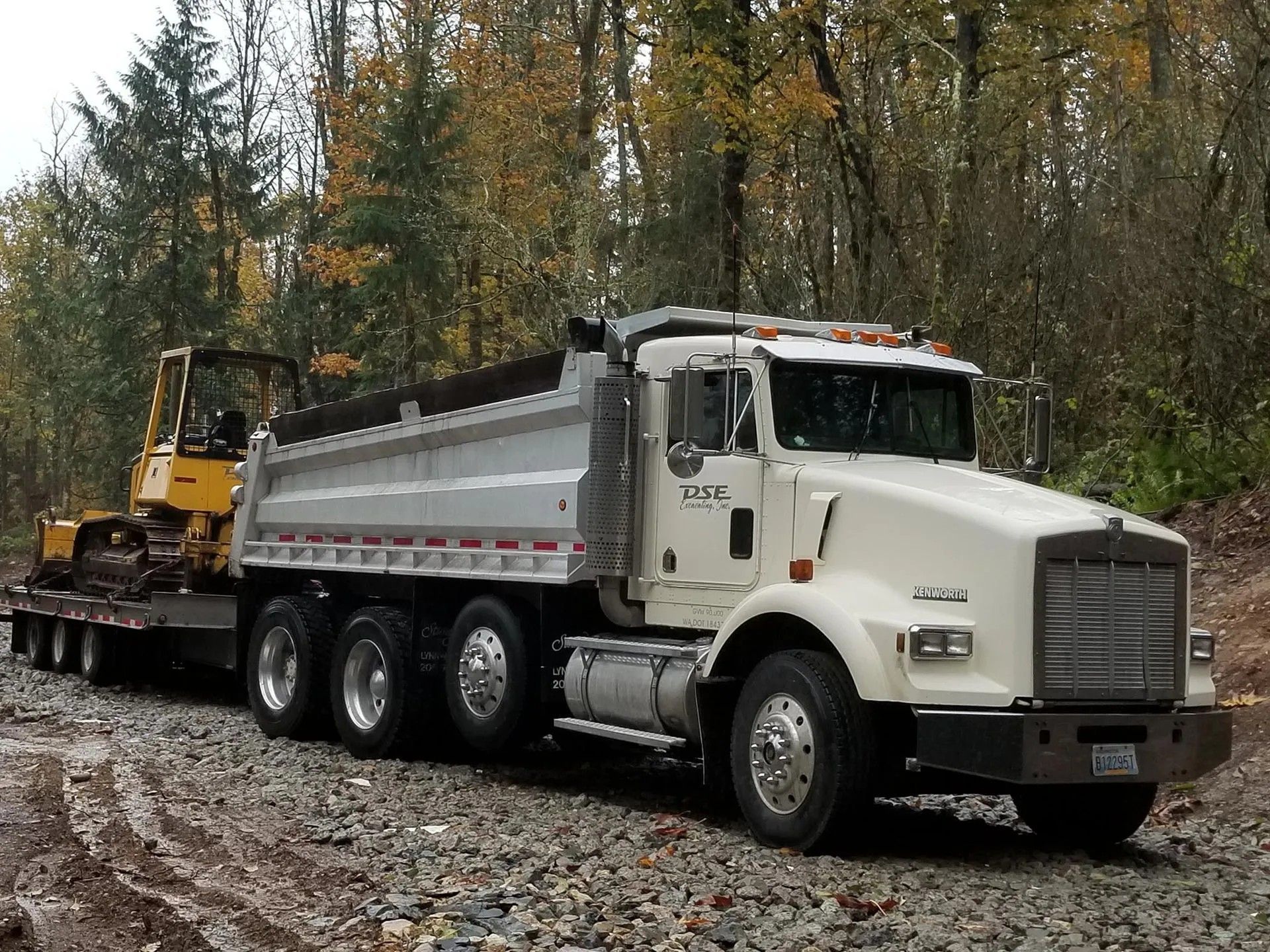 White dump truck hauling a bulldozer on a trailer on a gravel road, forest in background.