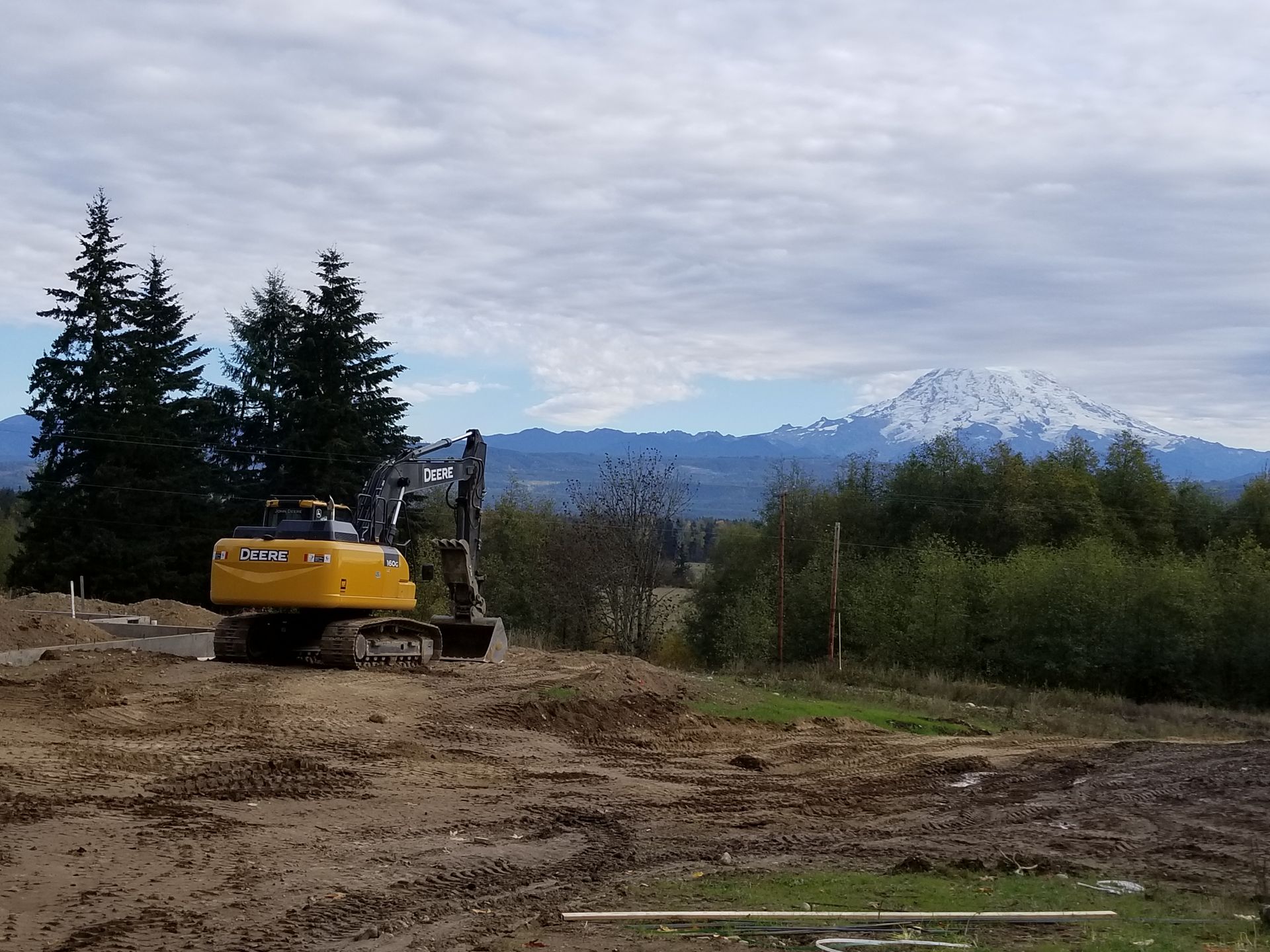 Yellow excavator on a muddy lot with a mountain in the distance. Overcast sky.