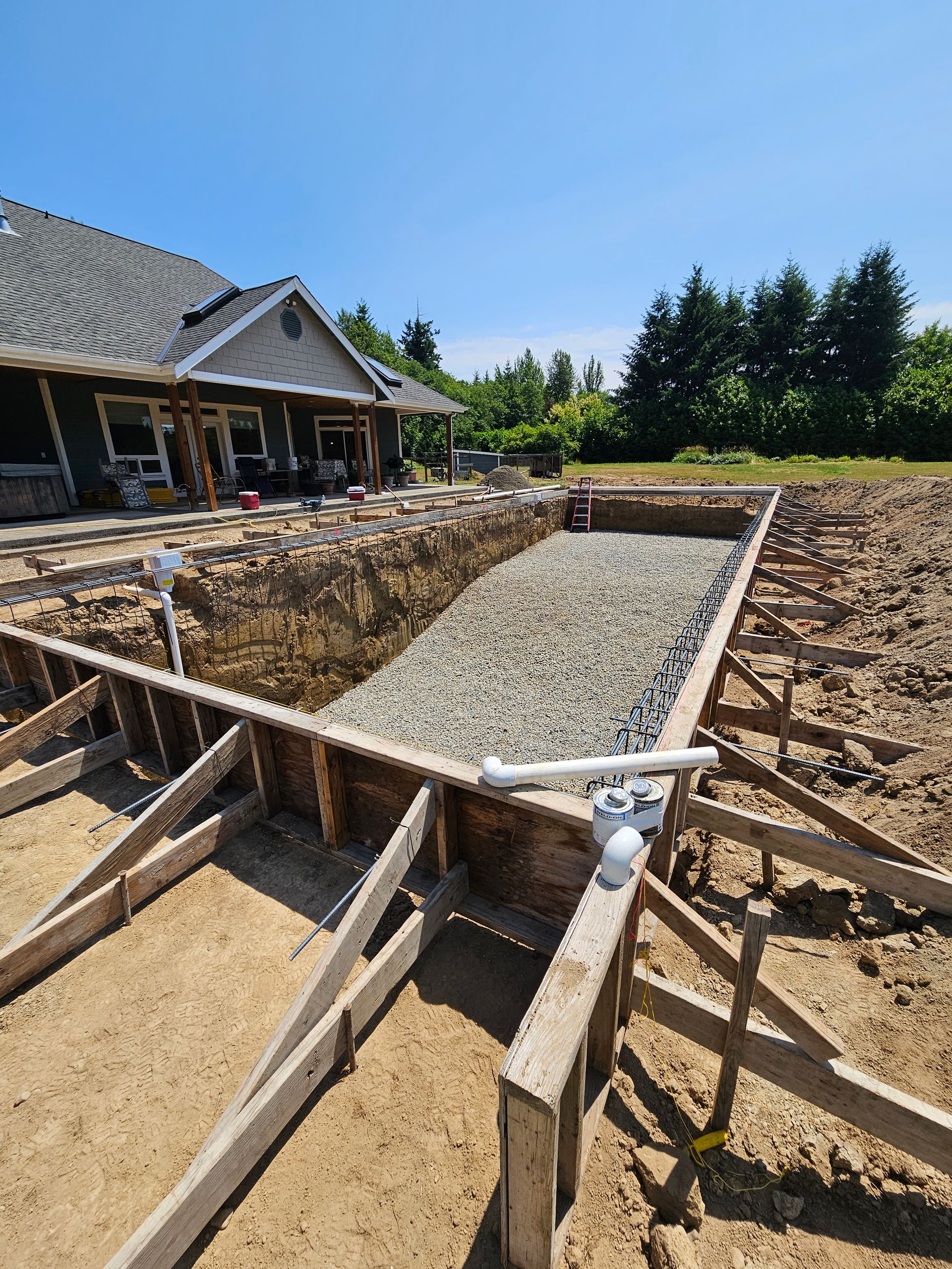 Construction site of a rectangular in-ground pool; gravel base, wooden formwork, house in background.