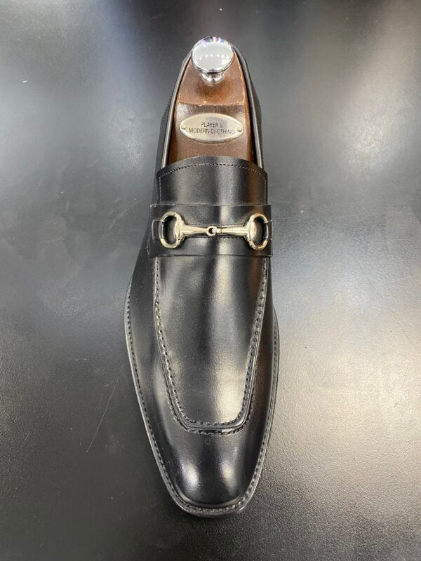 A pair of black loafers with a gold buckle on a black surface.