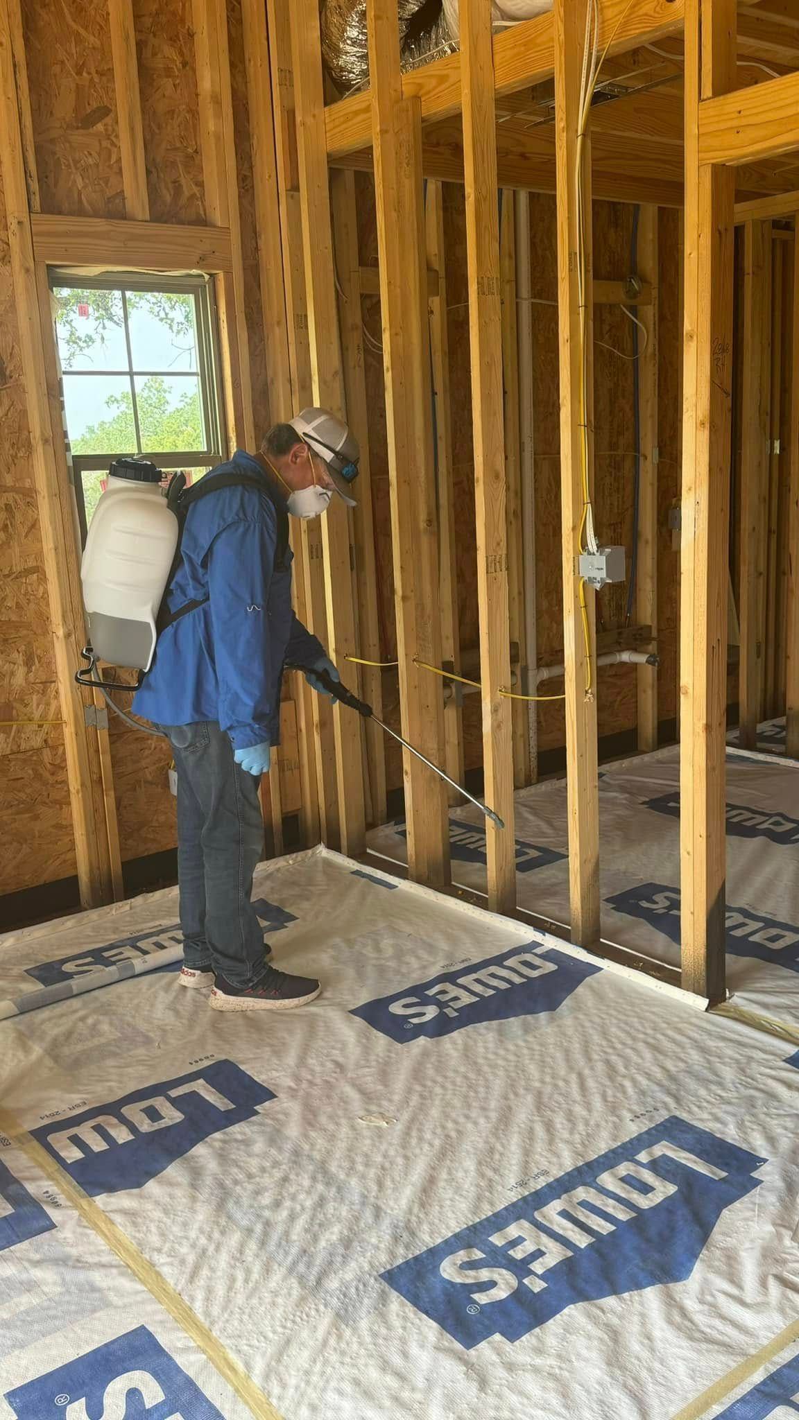 A person spraying insecticide in an unfinished house. Wearing a respirator, backpack sprayer, and gloves.