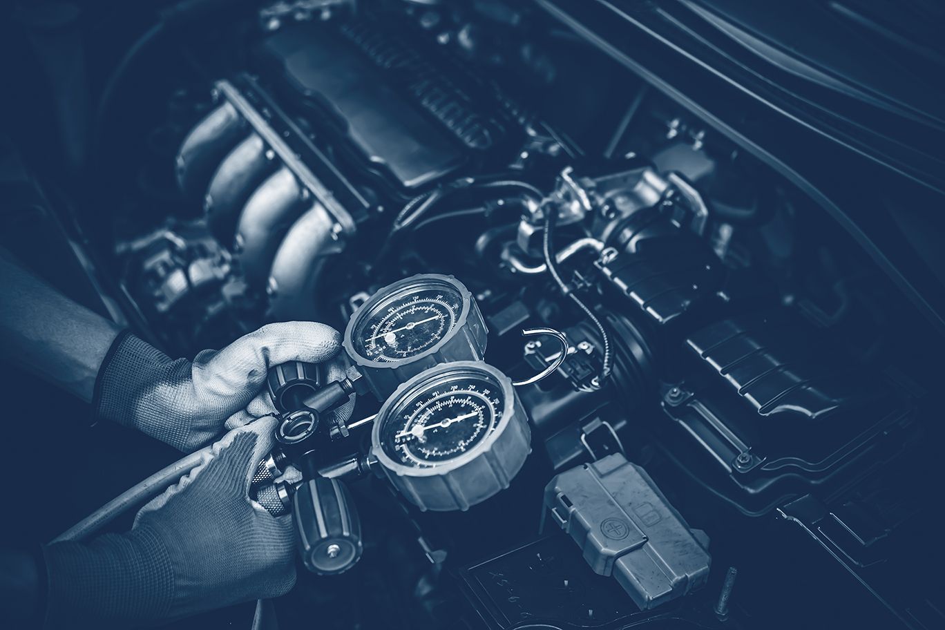 Mechanic using gauges to test a car engine, hands wearing gloves. Engine bay is dark blue.
