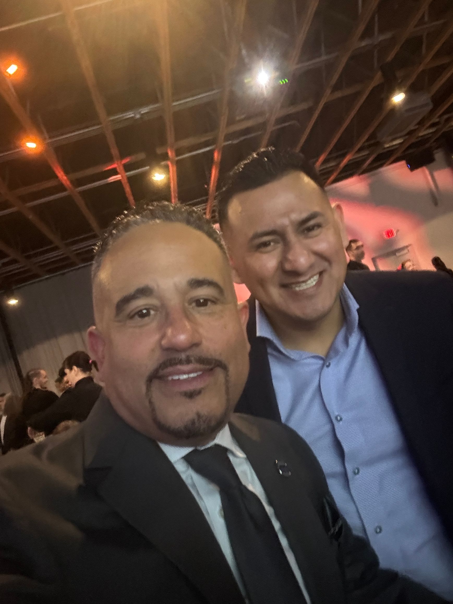 Two men smiling, close-up selfie; one in a suit and tie, the other in a button-up shirt and blazer, indoors.