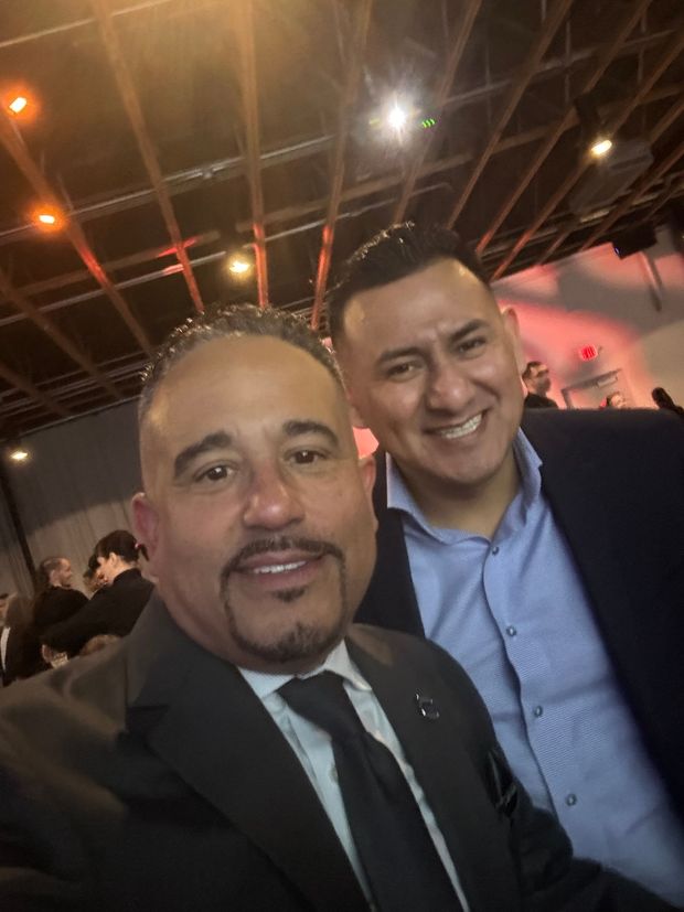 Two men smiling, close-up selfie; one in a suit and tie, the other in a button-up shirt and blazer, indoors.