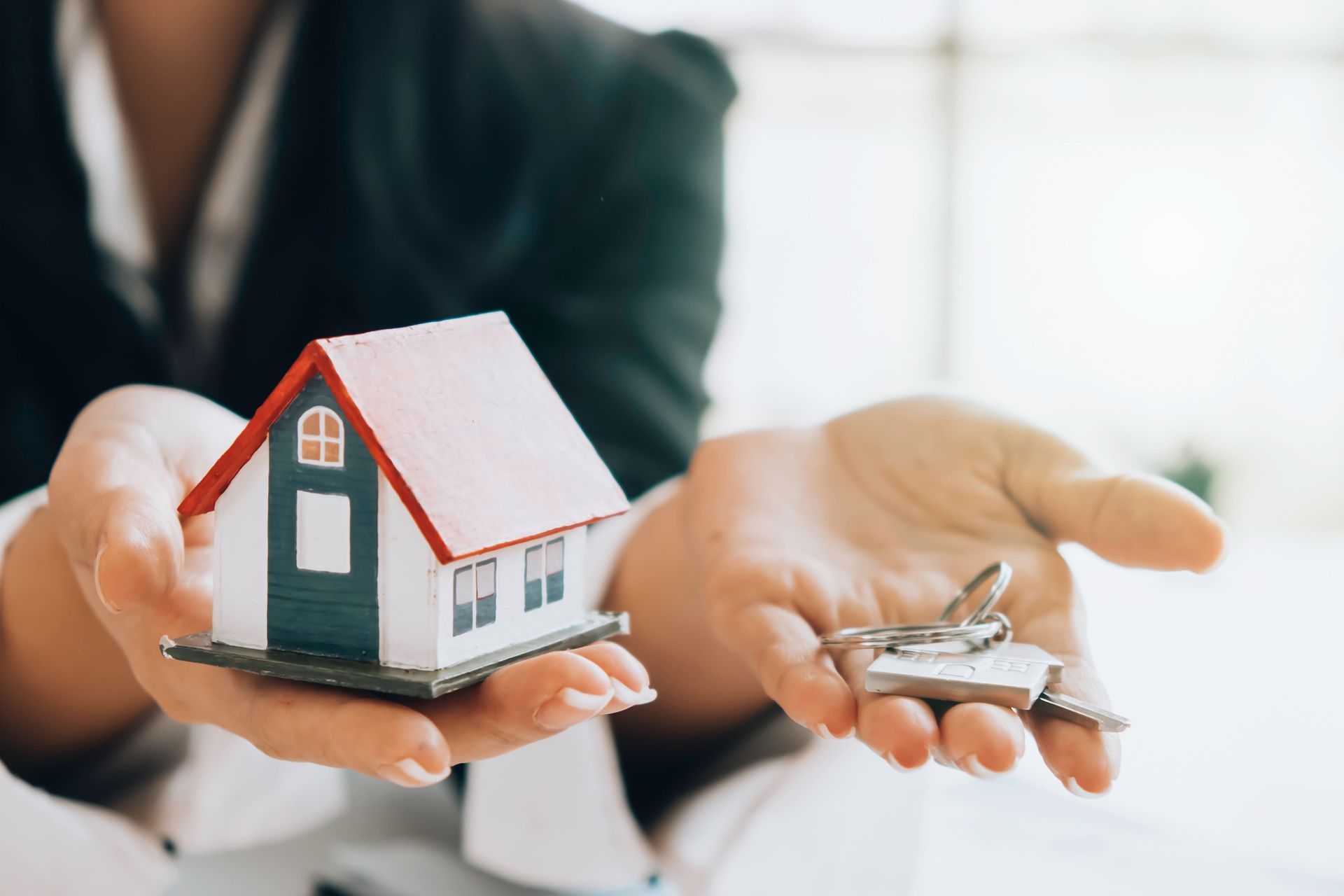 Person in business attire holding a miniature house and keys, symbolizing real estate.