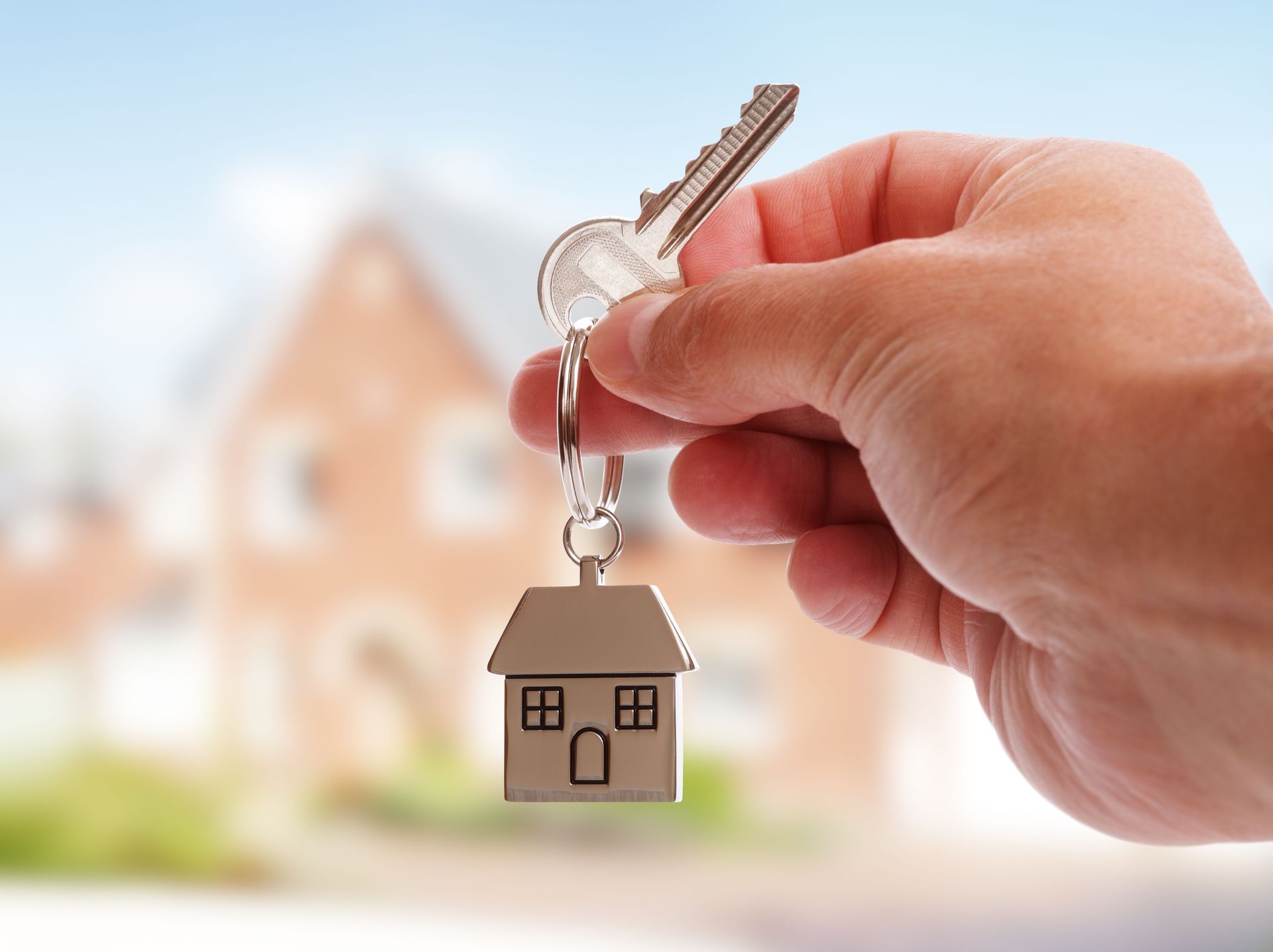 Hand holding house key with house-shaped keychain in front of a blurred house.