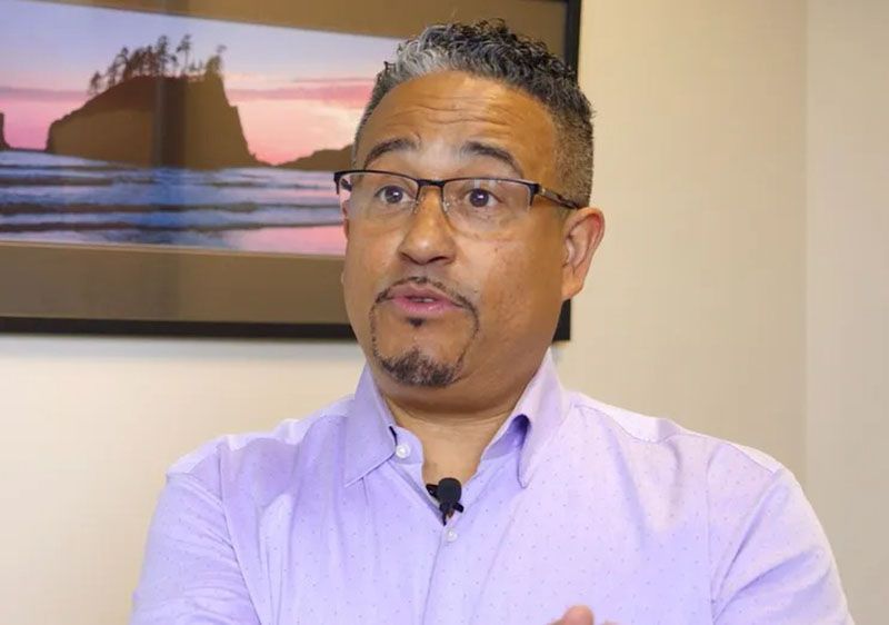 Man in purple shirt and glasses speaking in an office setting, with a landscape art in the background.